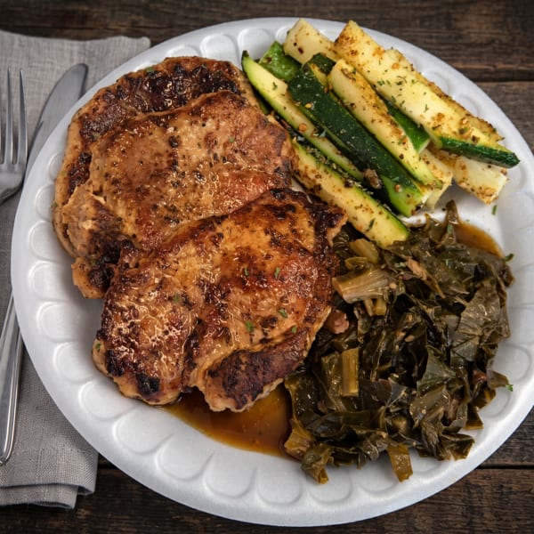 Three grilled pork chops, seasoned zucchini, and collard greens on a white plate with a fork and knife.