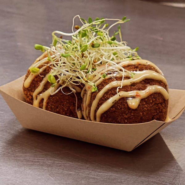 Two crispy brown arancini balls drizzled with sauce and topped with fresh sprouts in a paper tray.