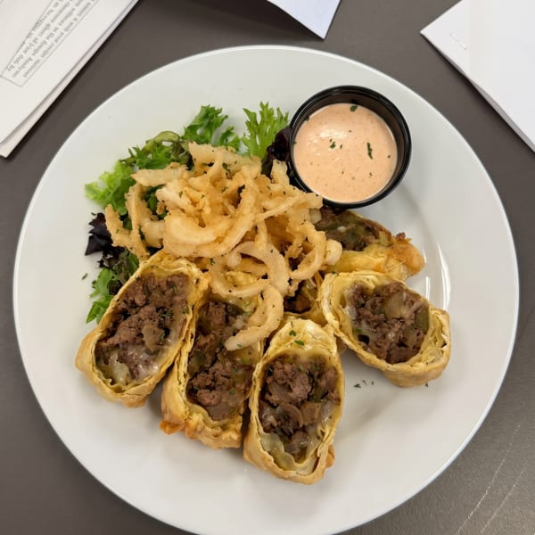 Cheesesteak egg rolls with crispy fried onions, a side salad, and dipping sauce on a white plate.