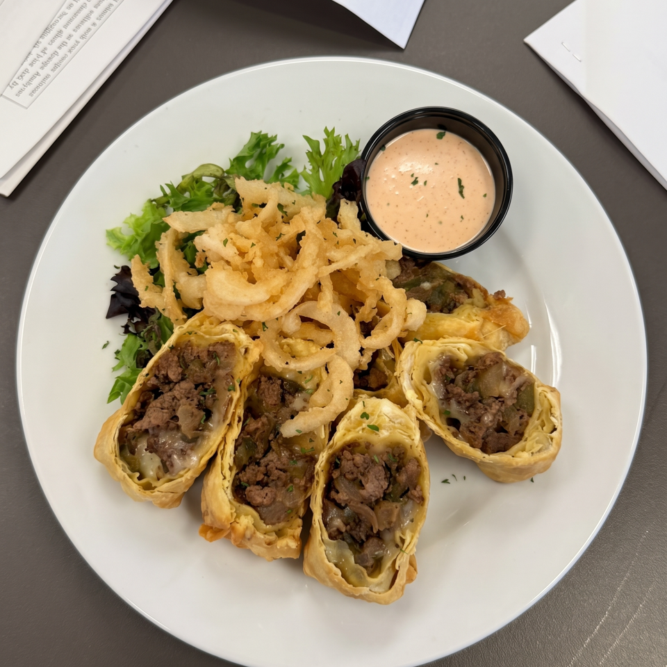 Cheesesteak egg rolls with crispy fried onions, a side salad, and dipping sauce on a white plate.