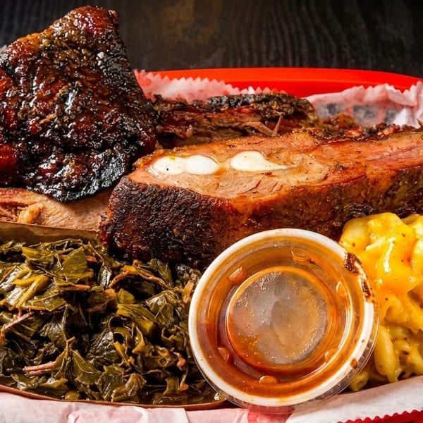 A red basket filled with barbecue ribs, pulled pork, collard greens, mac and cheese, and sauce.